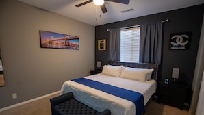 Un dormitorio con cama, mesita de noche, una silla, ventilador de techo, ventana con cortinas, un cuadro en la pared y el logotipo de Chanel.