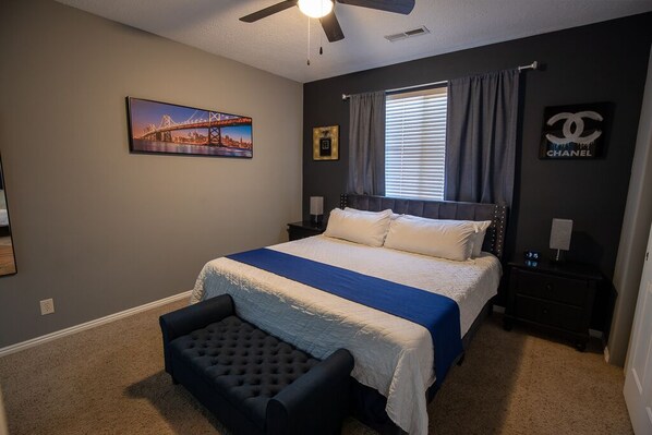 Um quarto com cama, mesa de cabeceira, cadeira, ventoinha no teto, janela com cortinas, quadro na parede e logotipo Chanel.