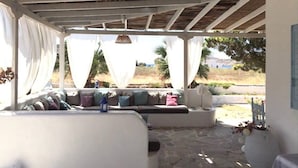 A covered outdoor seating area with a stone floor, white furniture, and draped white curtains. - Villa 100 M From THE Beach 6 Room IN IOS Island (Ios)
