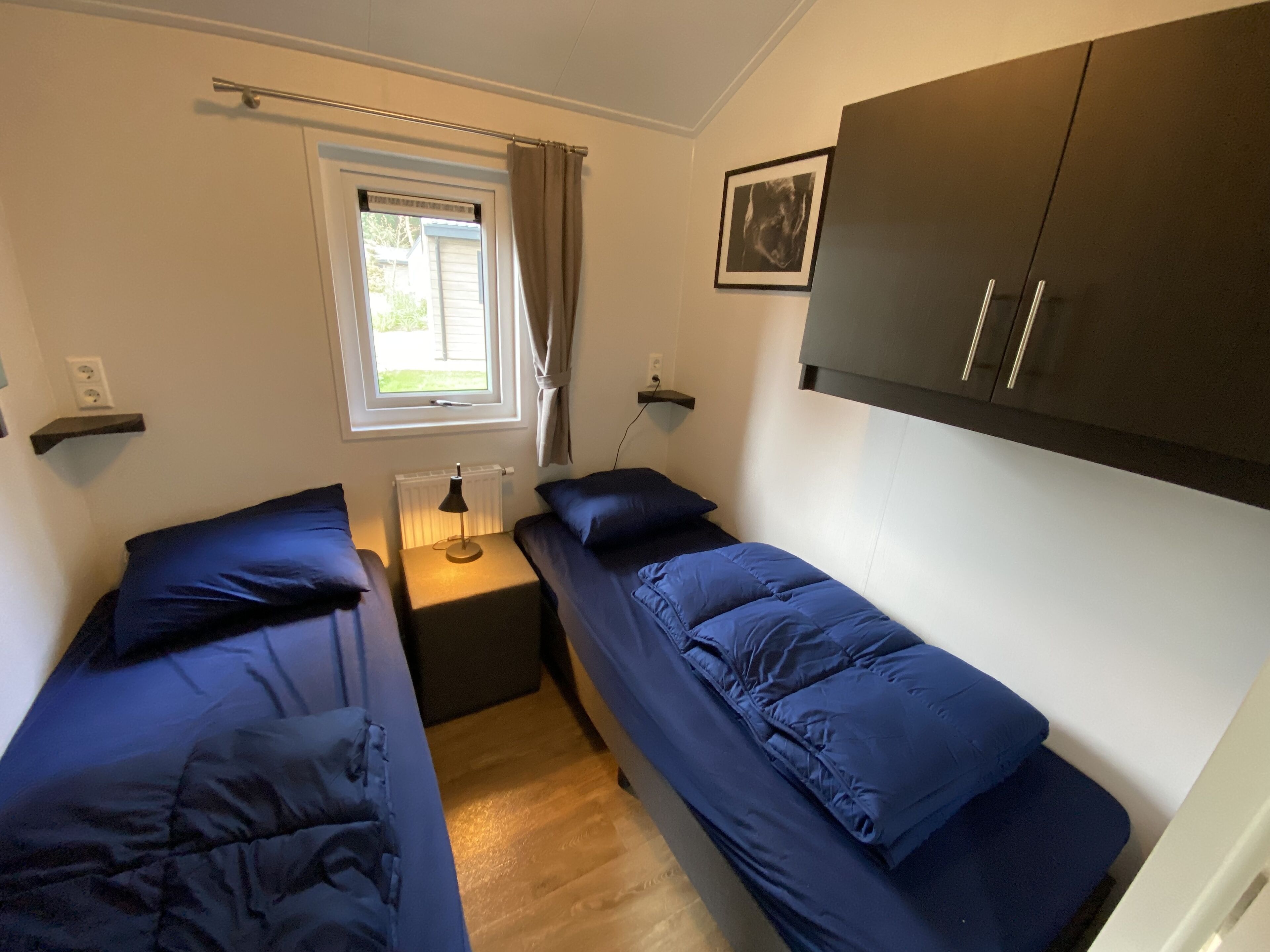 A small, well-lit room with two single beds, a window with curtains, a radiator, a wall-mounted shelf, and a framed picture.