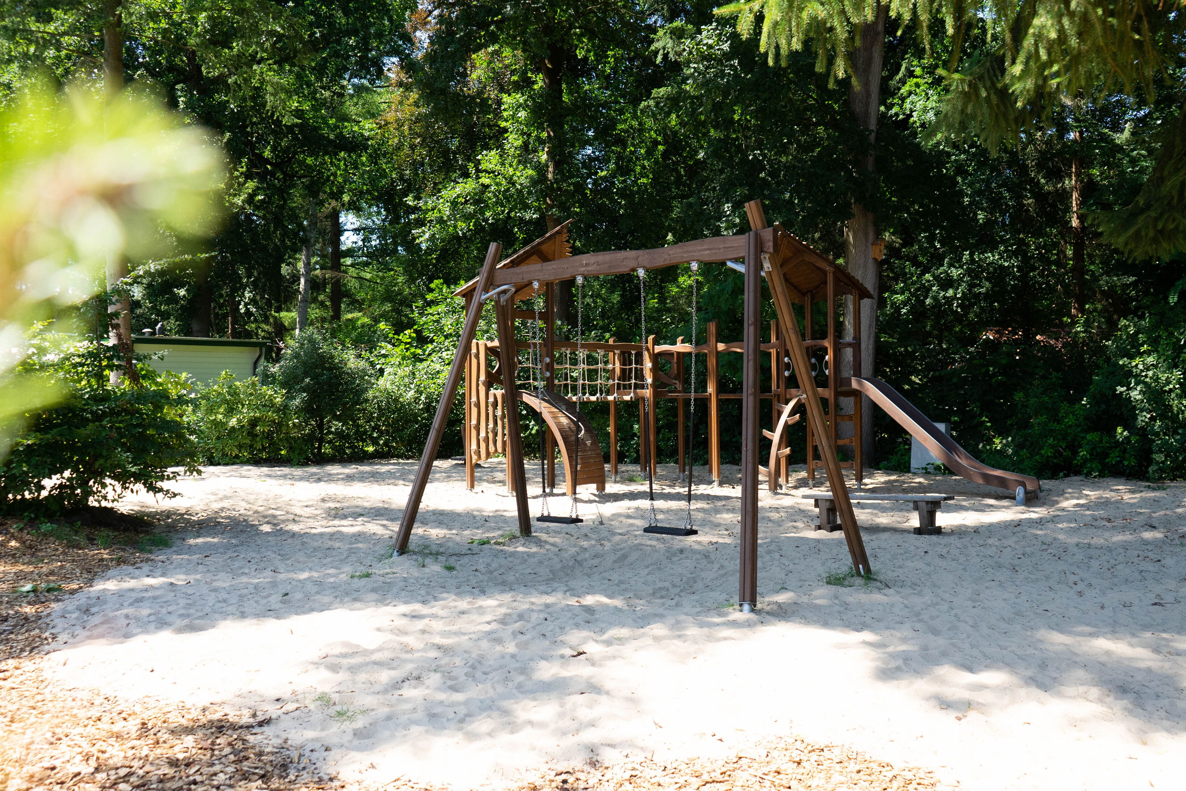 Een speelplaats met schommels en een glijbaan, omgeven door bomen en zand.