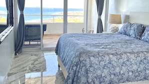 Quarto com cama grande, ventilador de teto e vista para o oceano por uma porta de vidro deslizante.