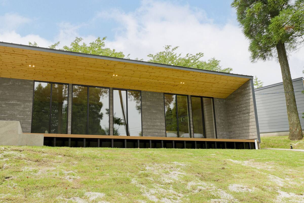 A modern building with large windows, a wooden sloped roof, and a grassy area in front.