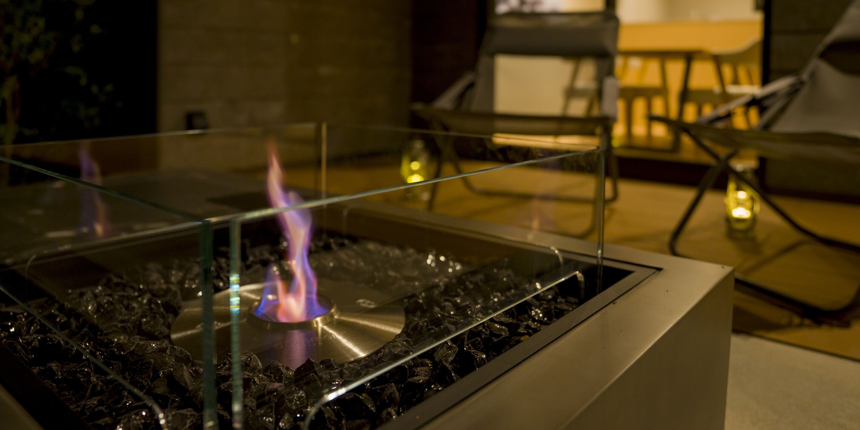A glass-top table with a central flame, surrounded by chairs and a table in a dimly lit room.