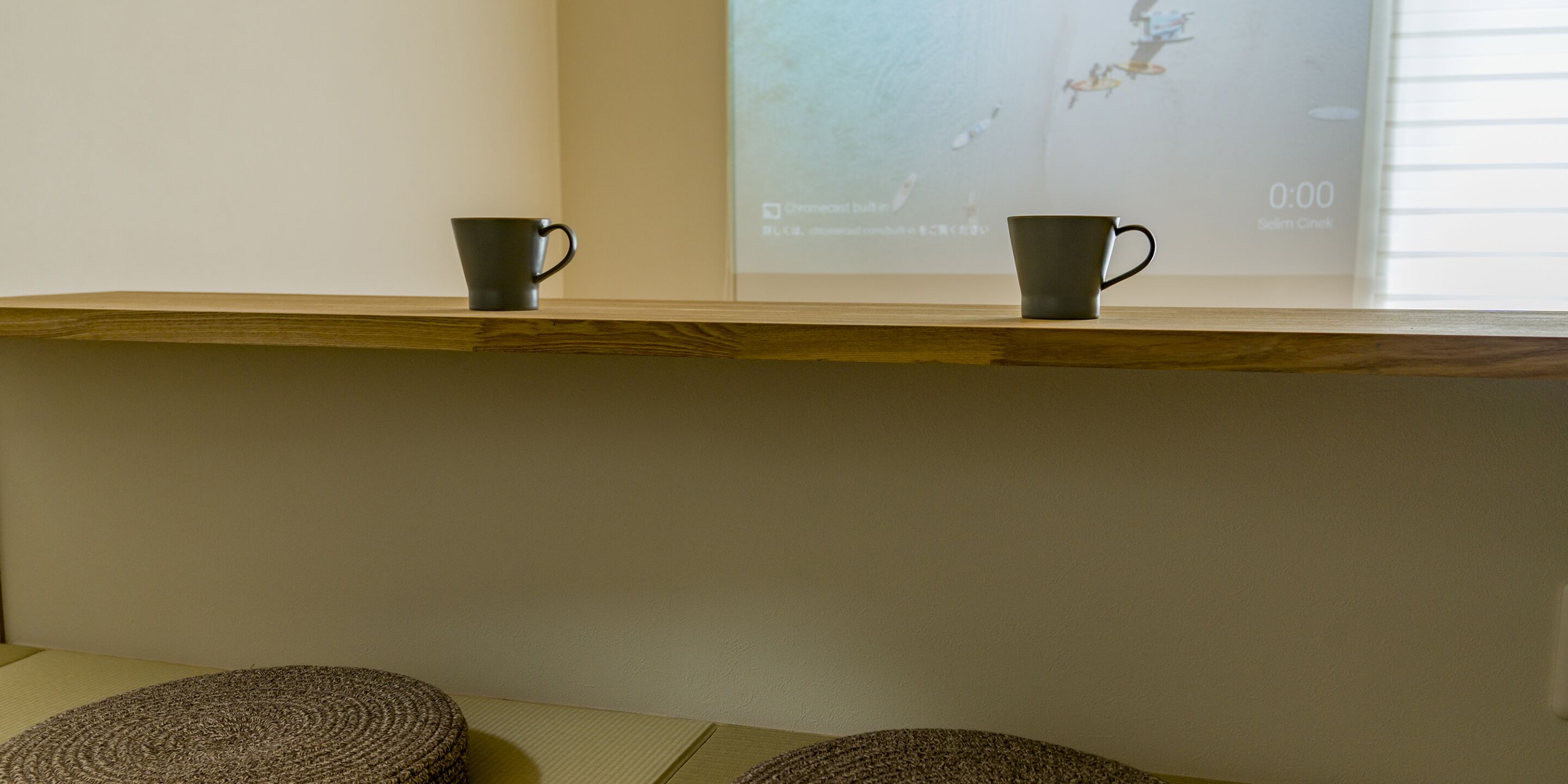 A wooden shelf with two dark mugs on it, a woven mat on the floor, and a projection screen in the background.