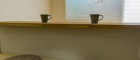 A wooden shelf with two dark mugs on it, a woven mat on the floor, and a projection screen in the background.