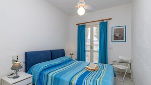 Une chambre à coucher comprenant un lit, une table de chevet, une lampe de bureau, une chaise et un ventilateur de plafond.