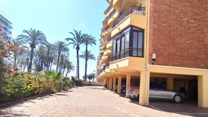 Un immeuble à plusieurs étages, doté d’une façade jaune, d’un parking souterrain et d’une allée pavée bordée de palmiers.