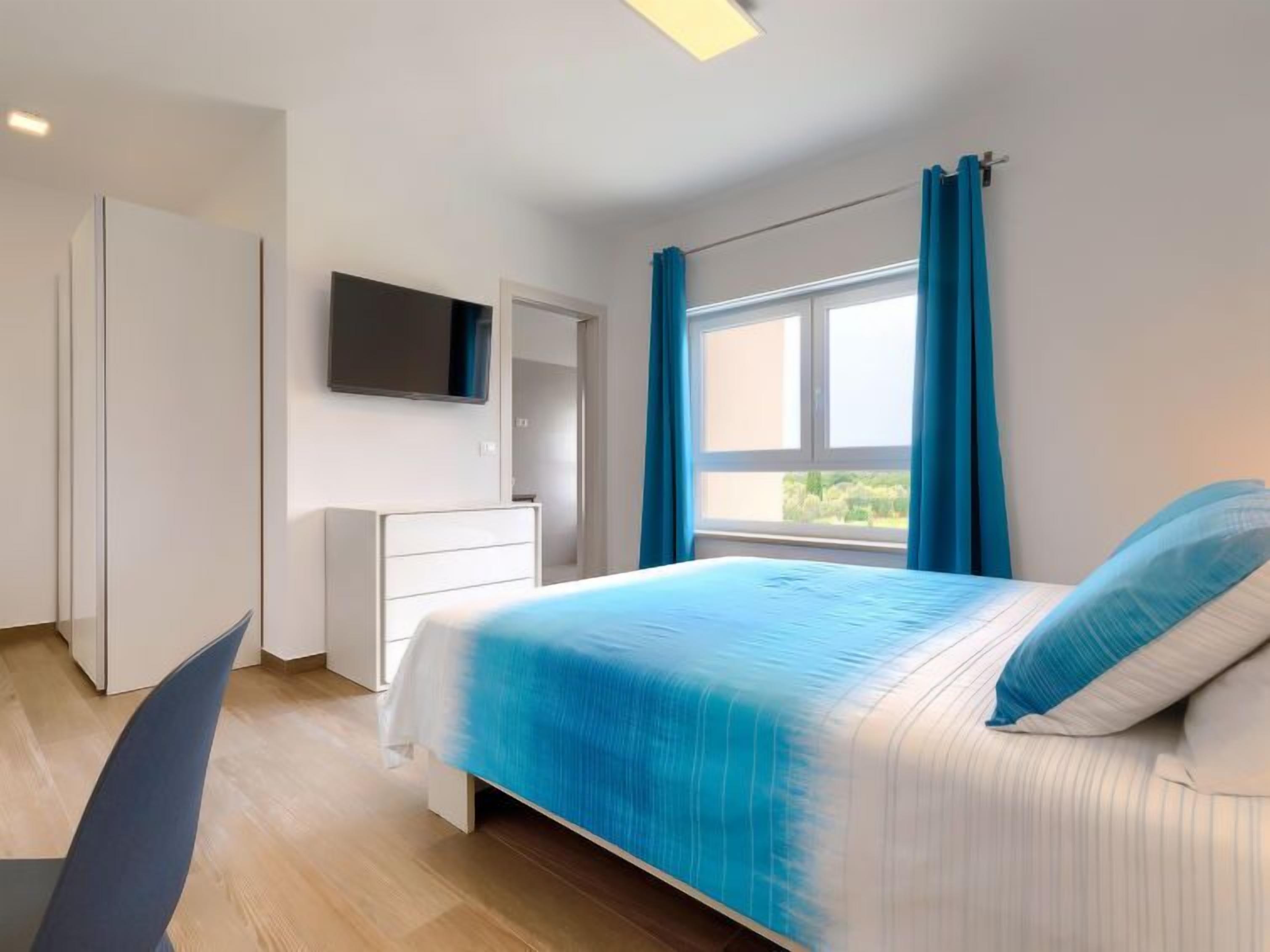 Un dormitorio con una cama, un televisor montado en la pared, un armario y una ventana con cortinas.