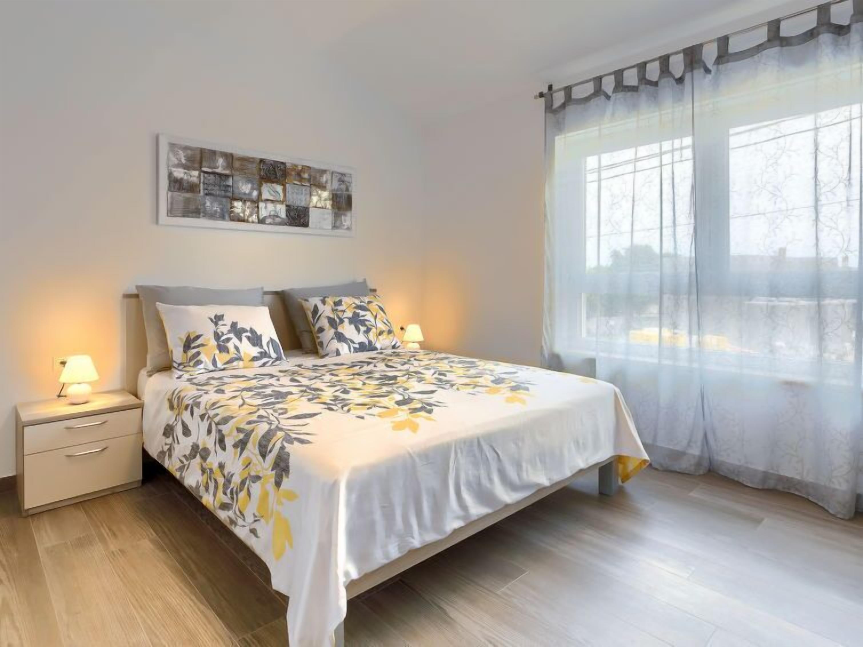 A bedroom with a bed, nightstands, a lamp, a window with sheer curtains, and a wall-mounted artwork.
