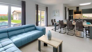 Un salon moderne avec un canapé bleu, un coin repas comprenant une table et des chaises, et une cuisine dotée de meubles sombres et d’une grande fenêtre.