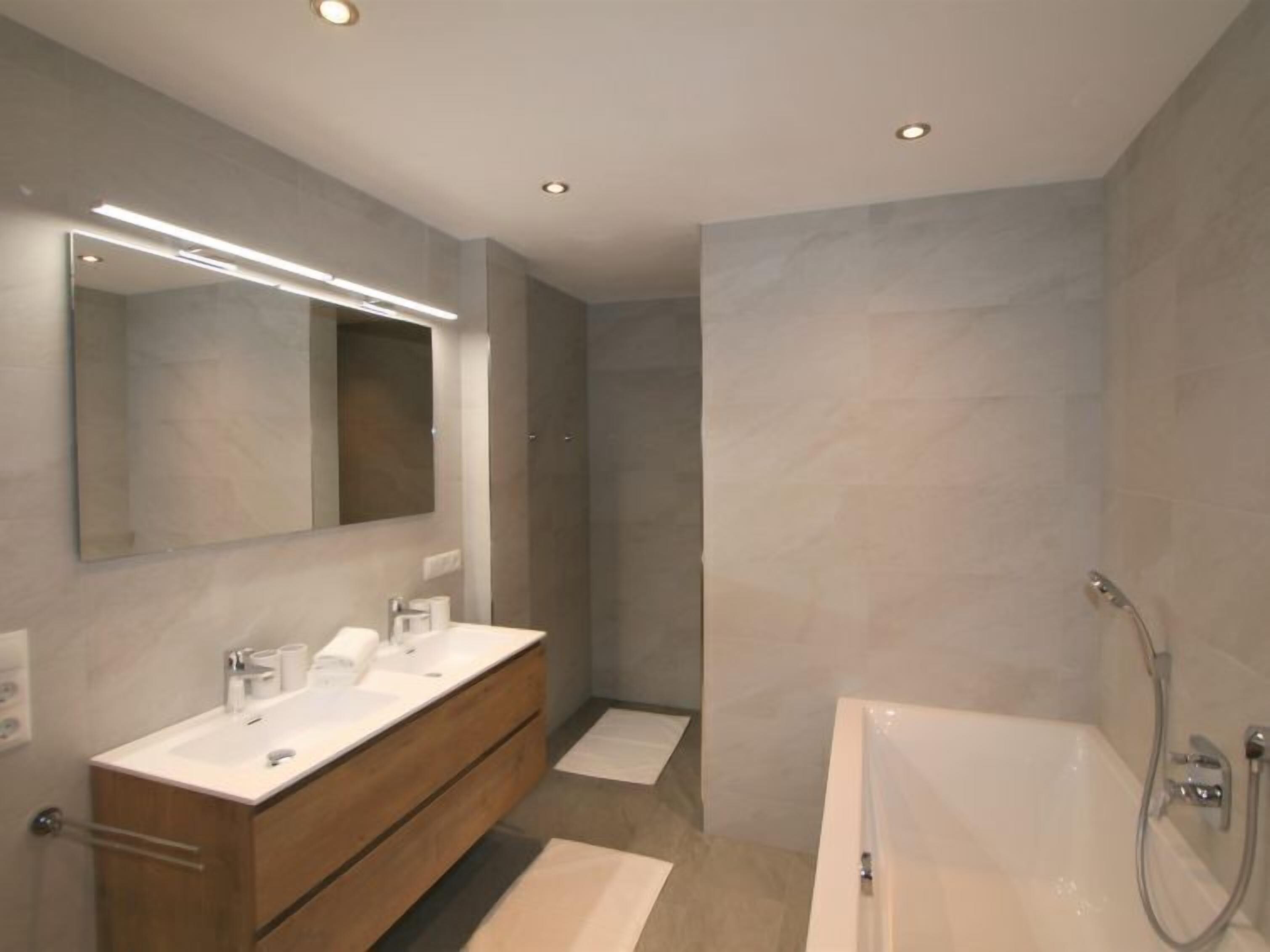 Un baño moderno con un espejo grande, dos lavamanos, una bañera e iluminación empotrada.