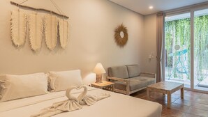 A hotel room with a bed, a sofa, a small table, a lamp, and a view of greenery through the window. - Malam Hari ApartHotel (Canggu)