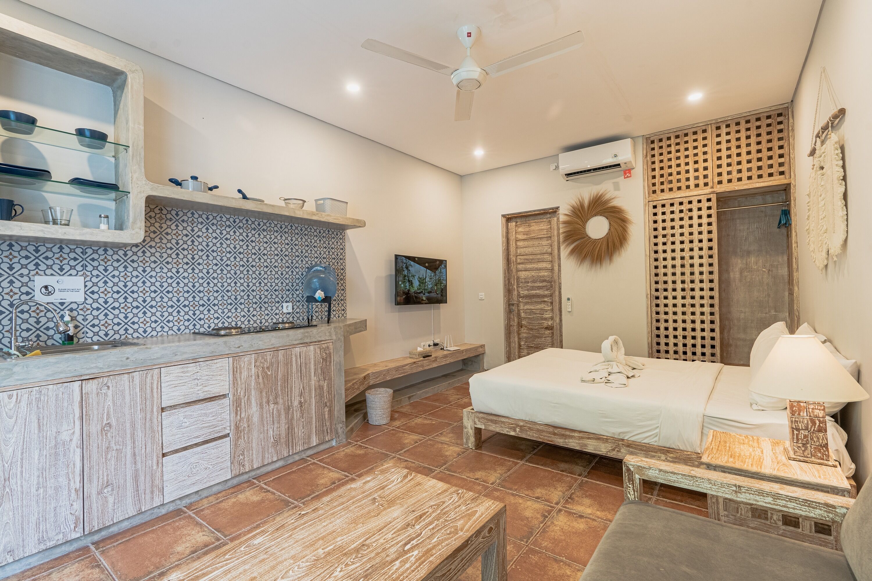 A compact hotel room with a kitchenette, a bed, a bedside table, a lamp, and a decorative wall piece.