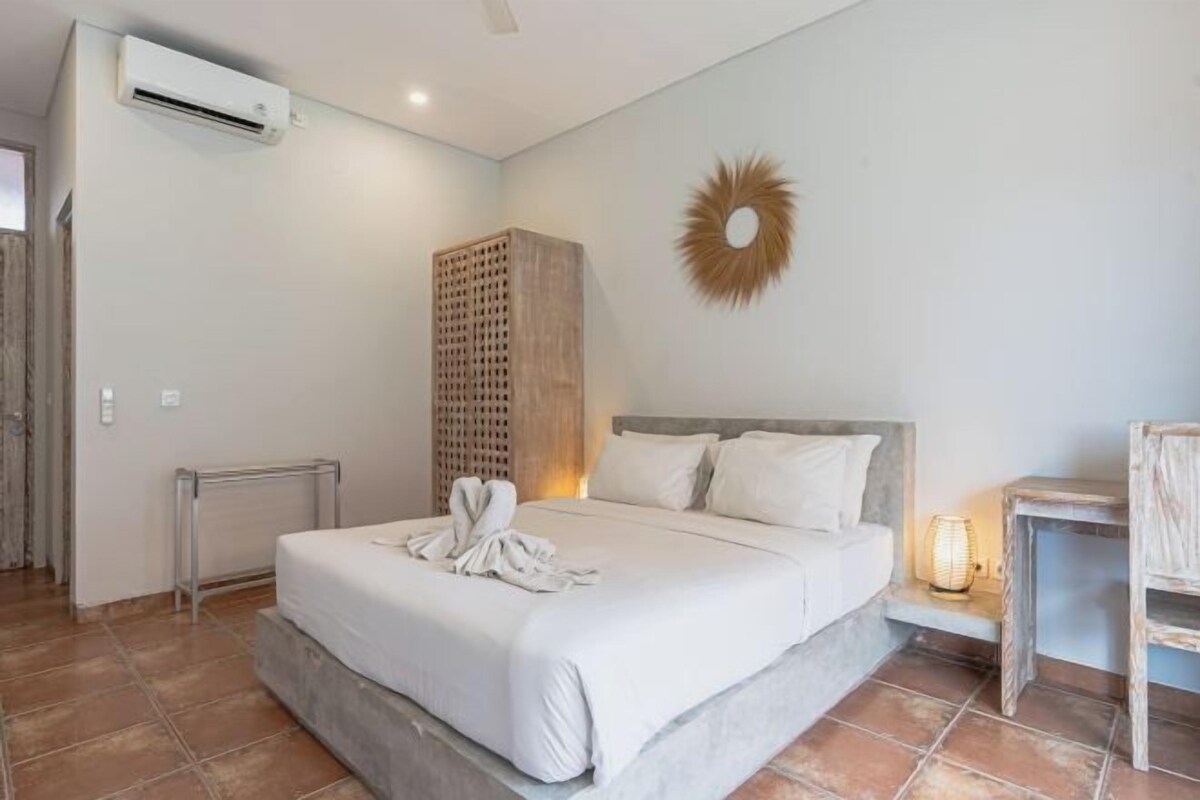 A hotel room with a bed, a radiator, a wall-mounted air conditioner, a decorative sunburst wall piece, and a small table with a lamp.
