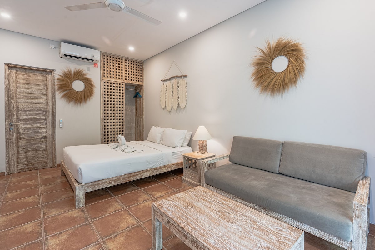 A bedroom with a bed, a grey sofa, a wooden coffee table, a wall-mounted air conditioner, and decorative wall hangings.