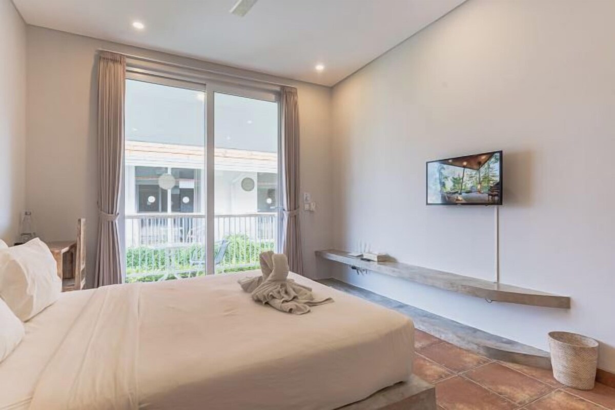 A hotel room with a bed, a TV mounted on the wall, a balcony with curtains, and a small shelf with items.