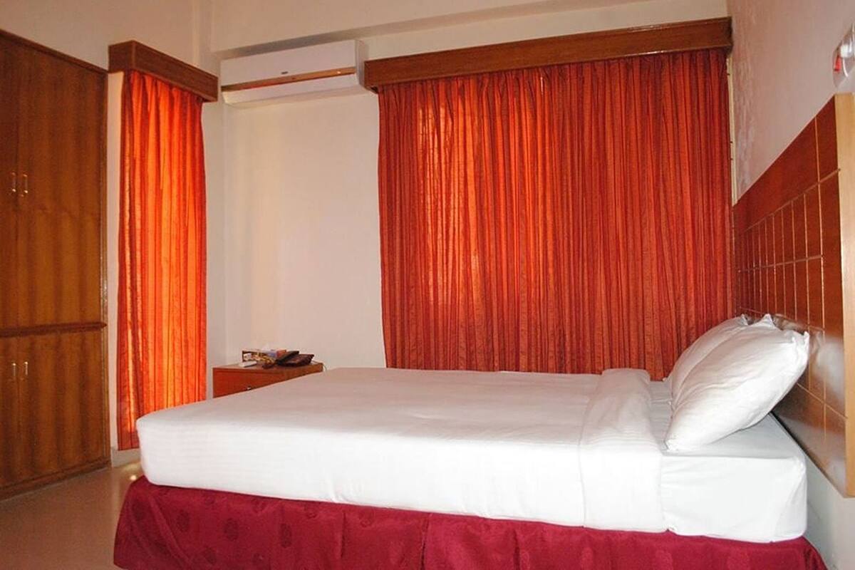 A hotel room with a bed, wooden wardrobe, and red curtains.