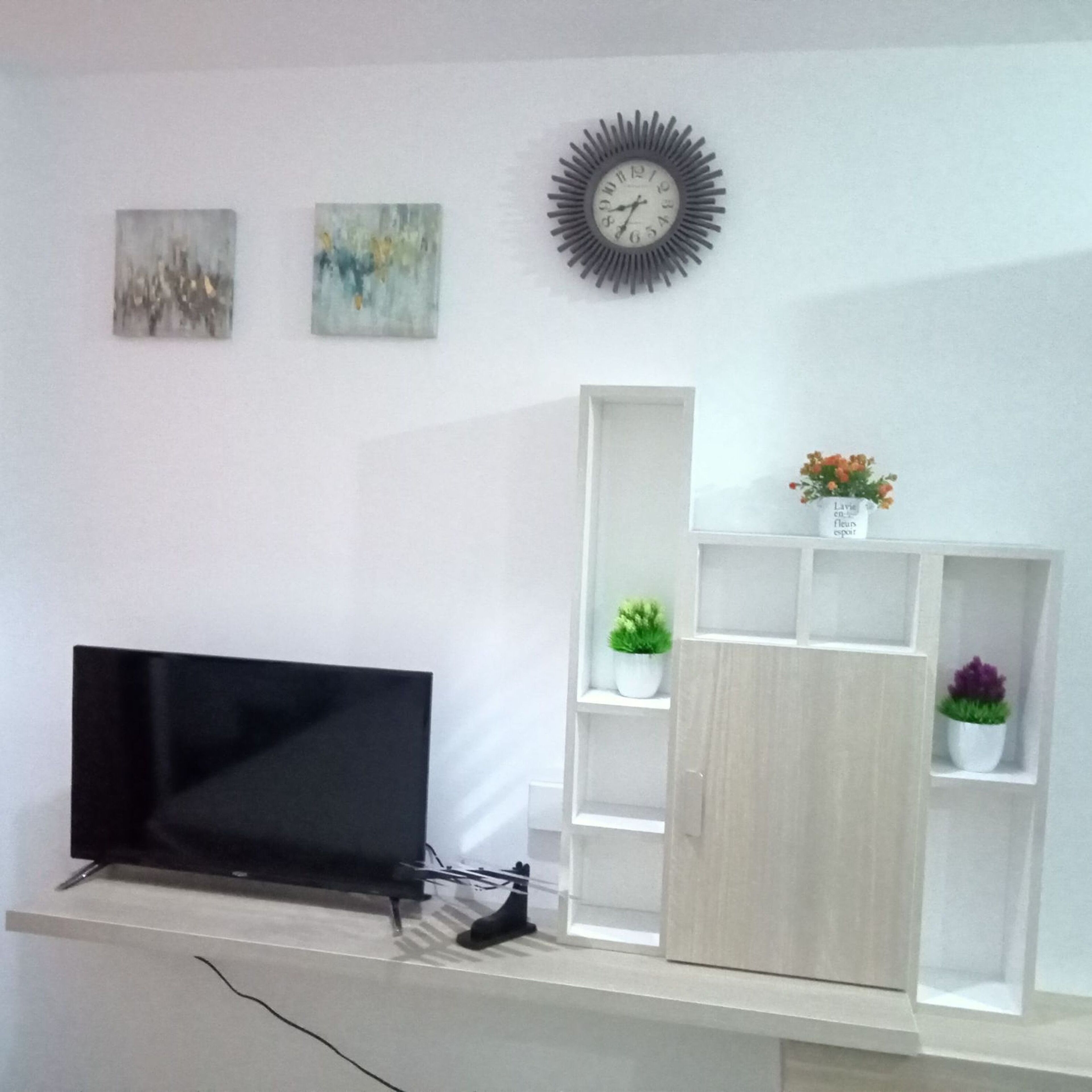 A modern living room with a flat-screen TV, a decorative clock, and a shelving unit with plants.