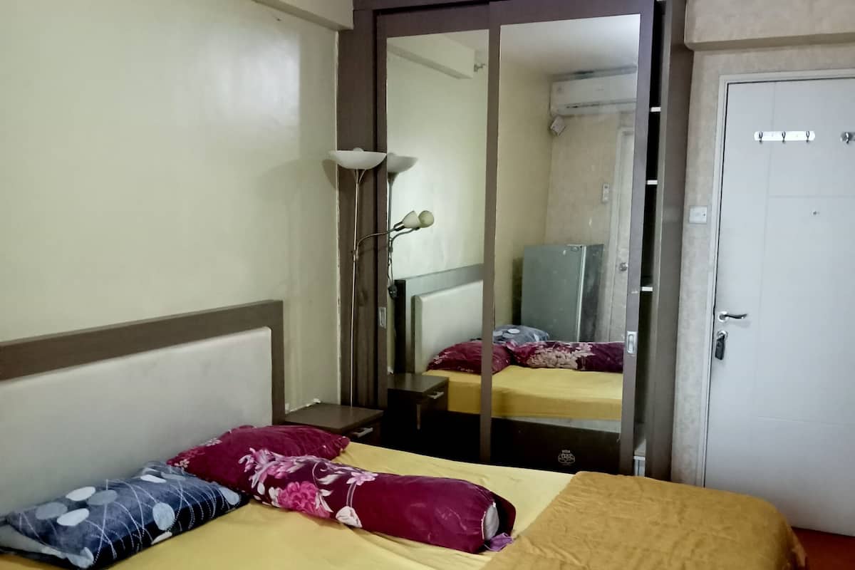A single bed with a yellow bedspread, a wooden headboard, and two pillows. There is a mirror on the wall, a white door, and a small table with a lamp.