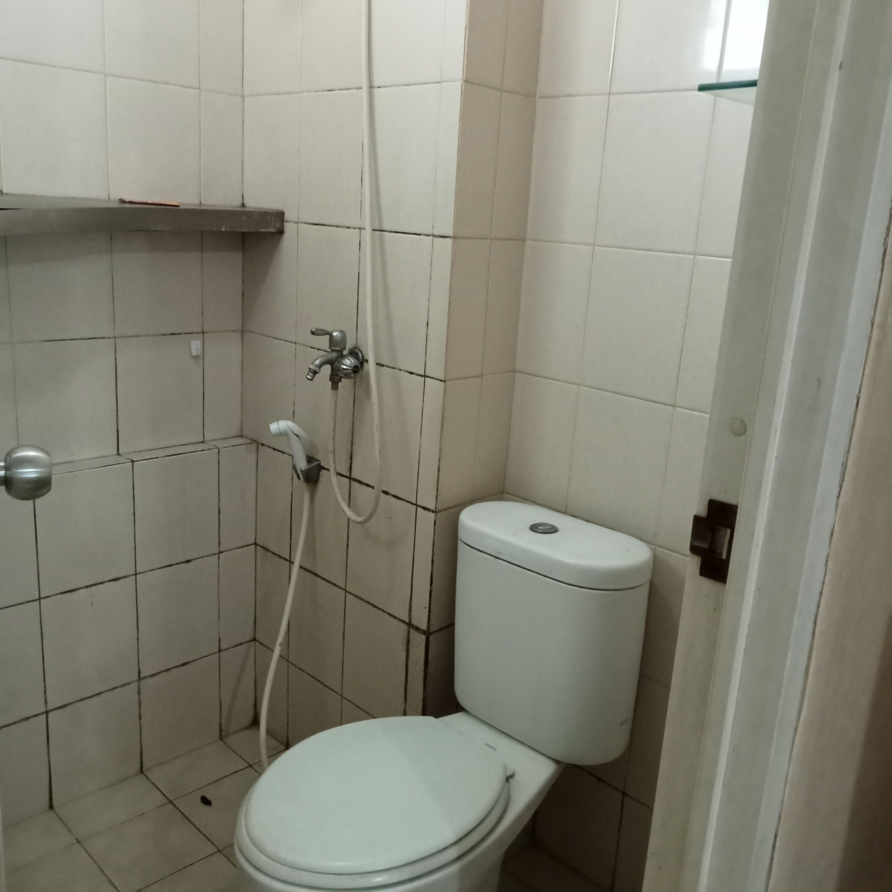A small, tiled bathroom with a toilet, a shower, and a shelf above the toilet.