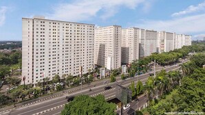 Une rangée d’immeubles résidentiels de grande hauteur, à plusieurs étages, une large route où circulent des véhicules, et de la verdure au premier plan.