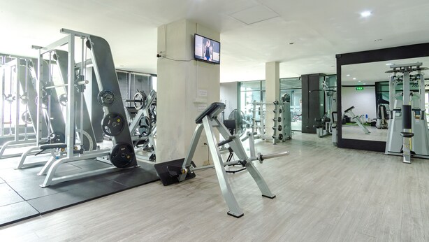 A gym with various exercise machines, a television mounted on the wall, and a large mirror reflecting the equipment. - Goodday Kathu (Kathu)