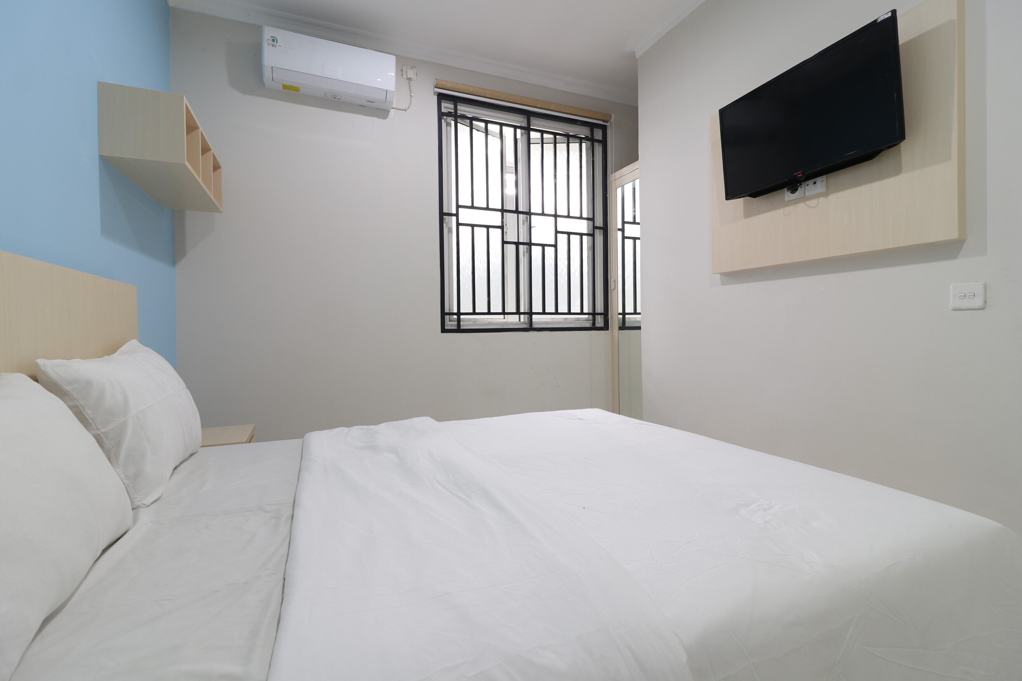 Habitación de hotel con cama, televisor montado en la pared, ventana con rejas y aire acondicionado.