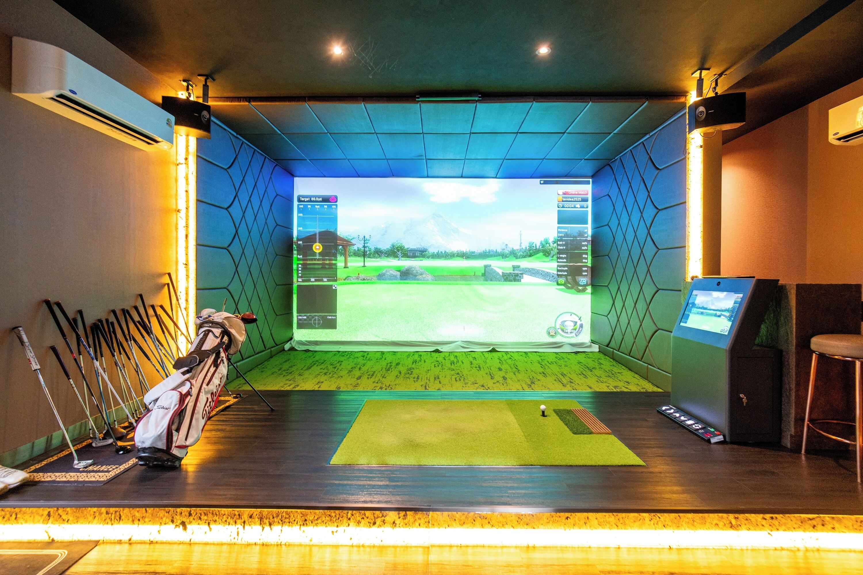 A golf simulator room with a large screen displaying a golf course, a golf bag, and several golf clubs.