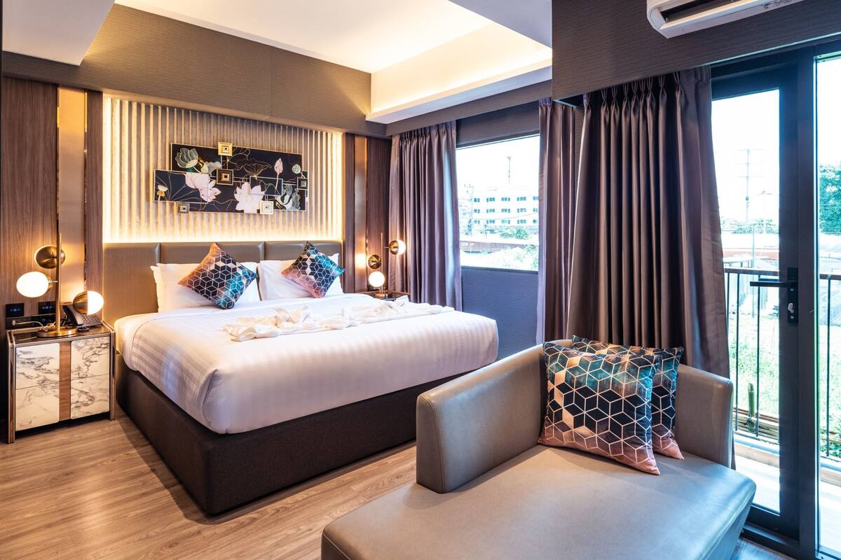 A modern hotel room with a large bed, bedside tables, a seating area, and a balcony with a view.