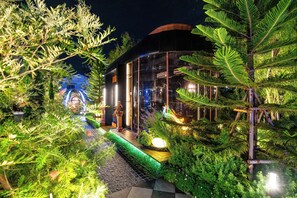 A modern building with a glass facade surrounded by lush greenery and a well-lit pathway. - The One Smart Living & Safety (Bangkok)