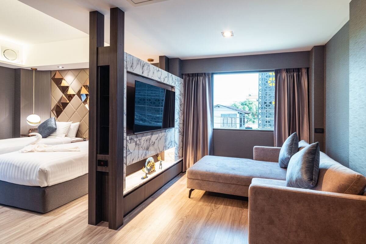 A modern hotel room with a bed, a sofa, a TV mounted on a marble wall, and a large window with curtains.