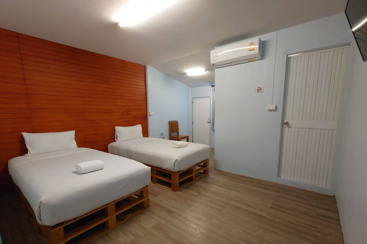 A hotel room with two beds, wooden flooring, a white door, and an air conditioning unit.