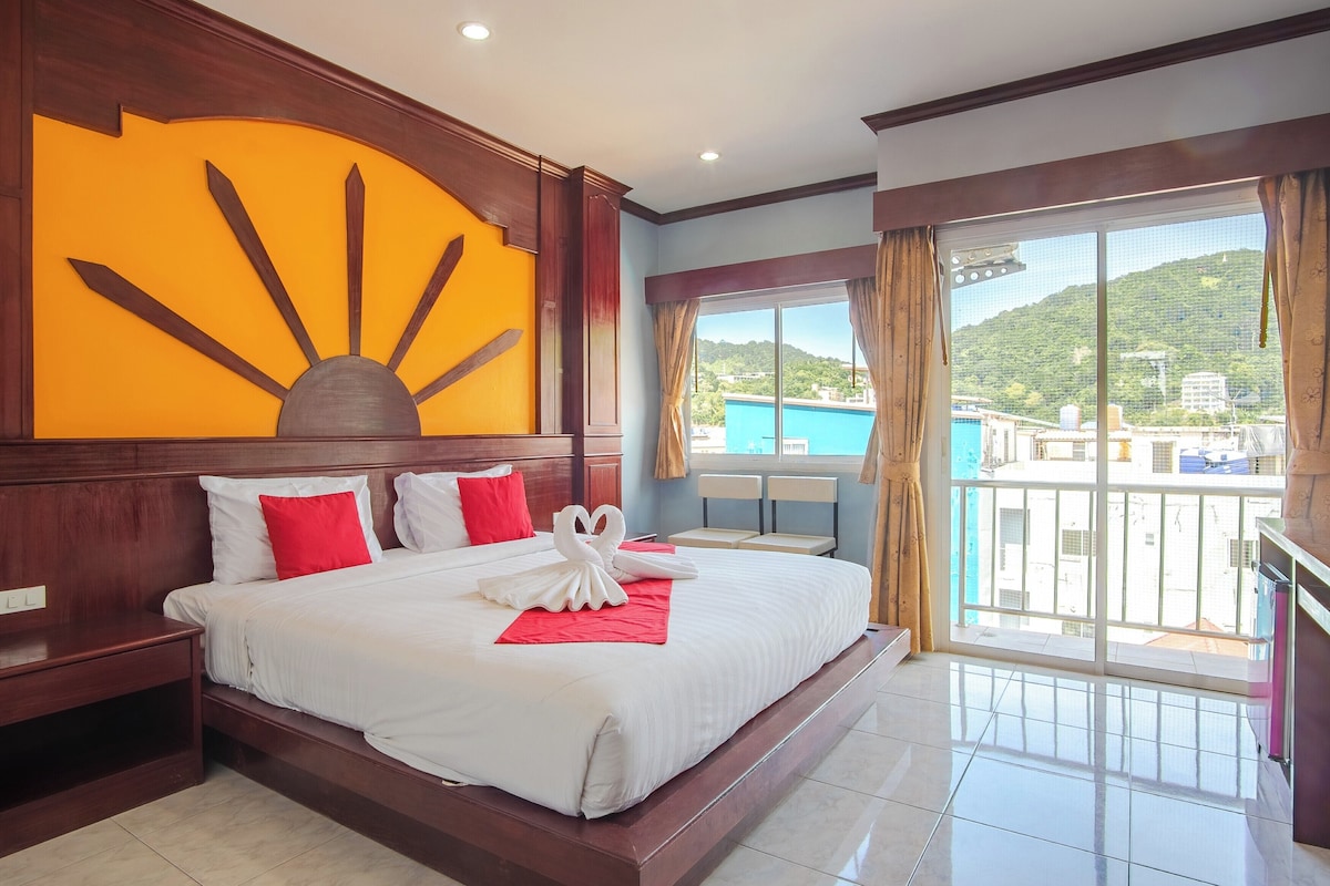 A hotel room with a large bed, a wooden headboard, a sun-shaped wall decoration, a balcony with a view of buildings and greenery, and a red pillow on the bed.