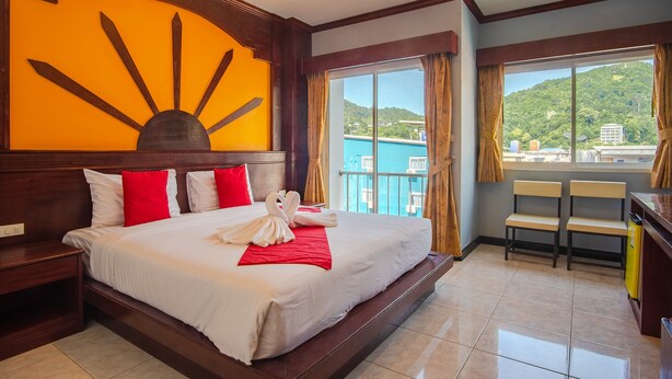 Une chambre d’hôtel avec un grand lit, une décoration murale en forme de soleil, un balcon avec vue sur la verdure et un bureau avec des chaises.