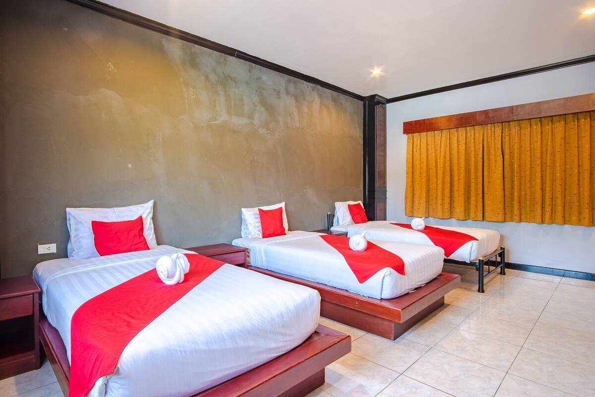 Een hotelkamer met drie bedden, elk voorzien van witte beddengoed en rode accenten, en een groot raam met gele gordijnen.