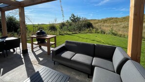 An outdoor patio with a wooden pergola, a black sofa, a table with a grill, and a view of a grassy field. - EuroParcs Texel (De Cocksdorp)