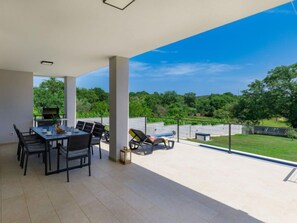 Un espacio comedor al aire libre cubierto, con mesa y sillas, una parrilla y vistas a un paisaje verde.