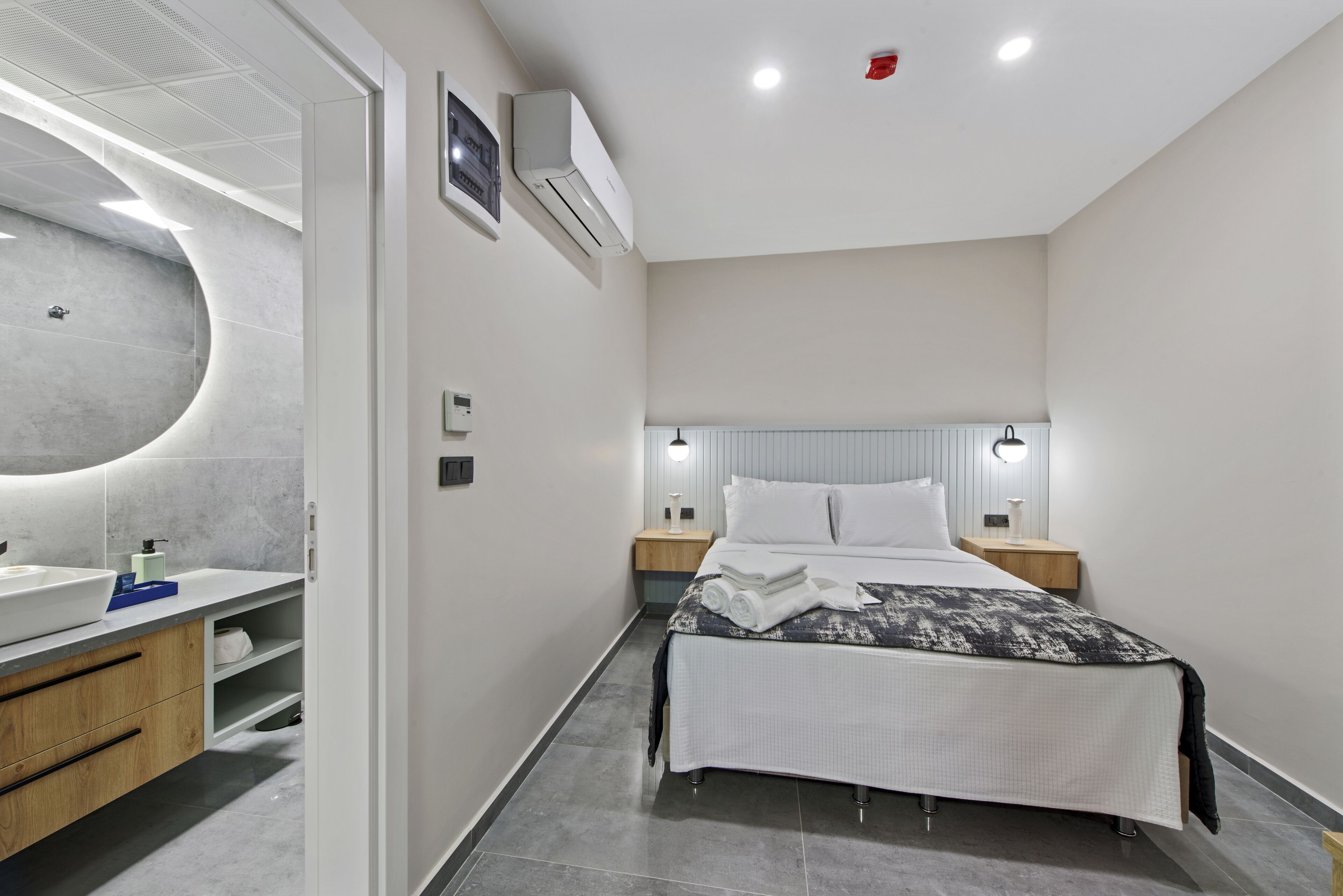 A modern hotel room with a curved wall, a large bathroom vanity, a bed with a white and black patterned bedspread, and two bedside tables with lamps.