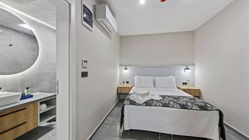 A modern hotel room with a curved wall, a large bathroom vanity, a bed with a white and black patterned bedspread, and two bedside tables with lamps.
