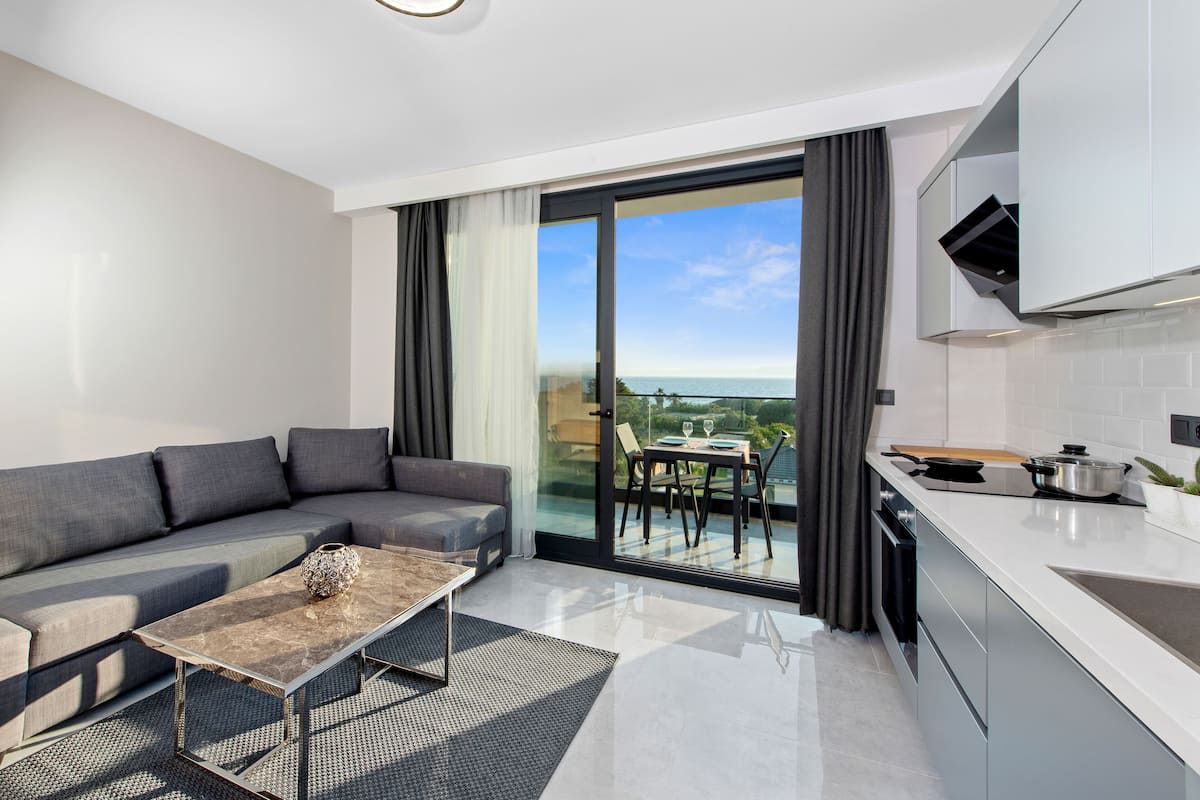 A modern living room with a sofa, a coffee table, and a dining area with a table and chairs. There is a kitchen area with a stove and cabinets. The room has large windows with a view of the ocean.
