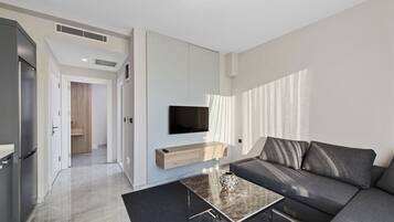Un salon moderne comprenant un canapé, une table basse, un téléviseur fixé au mur et un coin cuisine avec un réfrigérateur.