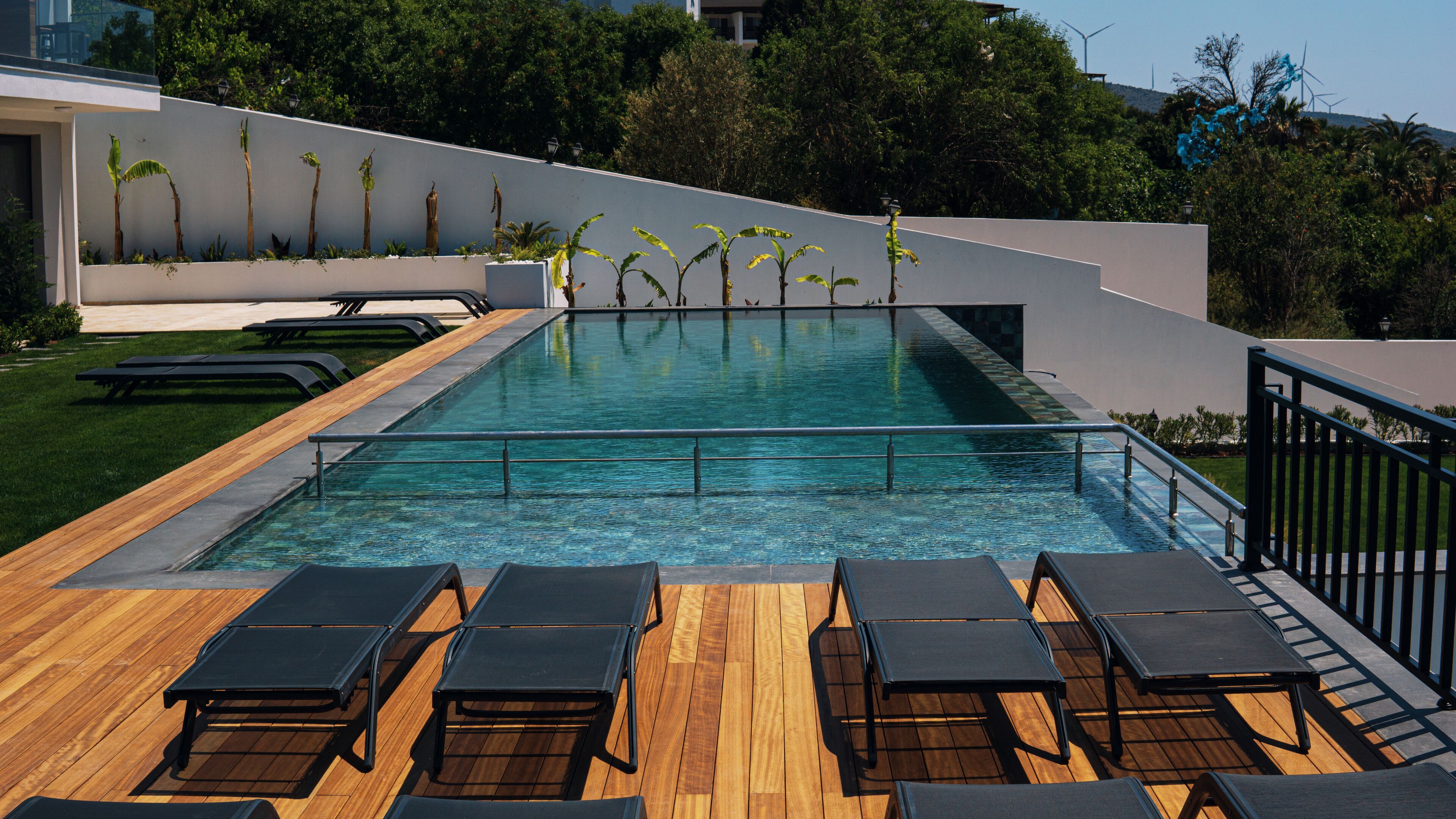 Piscina exterior moderna con terraza de madera y sillones de descanso, rodeada de vegetación y un muro blanco.
