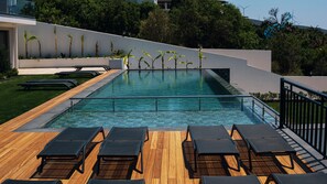 A modern outdoor pool with wooden deck and lounge chairs, surrounded by greenery and a white wall.