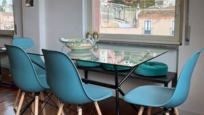 Un coin repas avec une table en verre et des chaises de couleur turquoise, une vue sur les bâtiments à travers une grande fenêtre, et un petit objet décoratif posé sur la table.