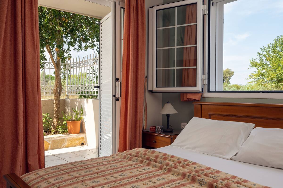 A bedroom with a bed, a window, curtains, a nightstand, a lamp, and a view of the outdoors.