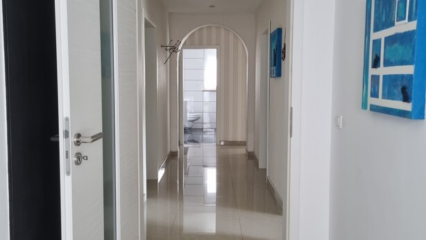 Un couloir au design moderne, avec un sol carrelé brillant et des murs blancs. Une porte se trouve à gauche et une œuvre d’art encadrée à droite.