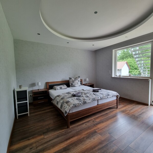 A bedroom with a wooden bed, a nightstand, a lamp, and a window with a view of a house and trees.