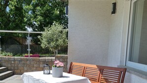 Une terrasse extérieure avec une table recouverte d’une nappe blanche, un banc en bois et des plantes en pot ornées de fleurs roses.