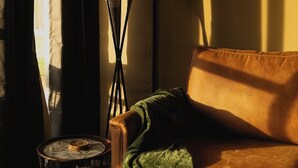 Ein Zimmer mit einer braunen Ledersofa, einer grünen Decke, einem kleinen runden Tisch mit einem weißen Gegenstand darauf und einer hohen Stehlampe.